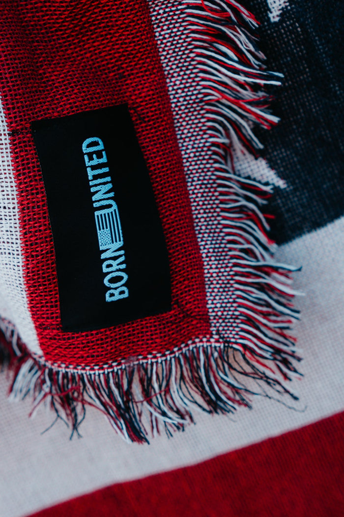 Close-up of the American Flag Jacquard Blanket featuring red, white, and blue resembling Old Glory. A black tag reads BORN UNITED. The fringe adds texture to the edge. Brand: Born United Private Label.