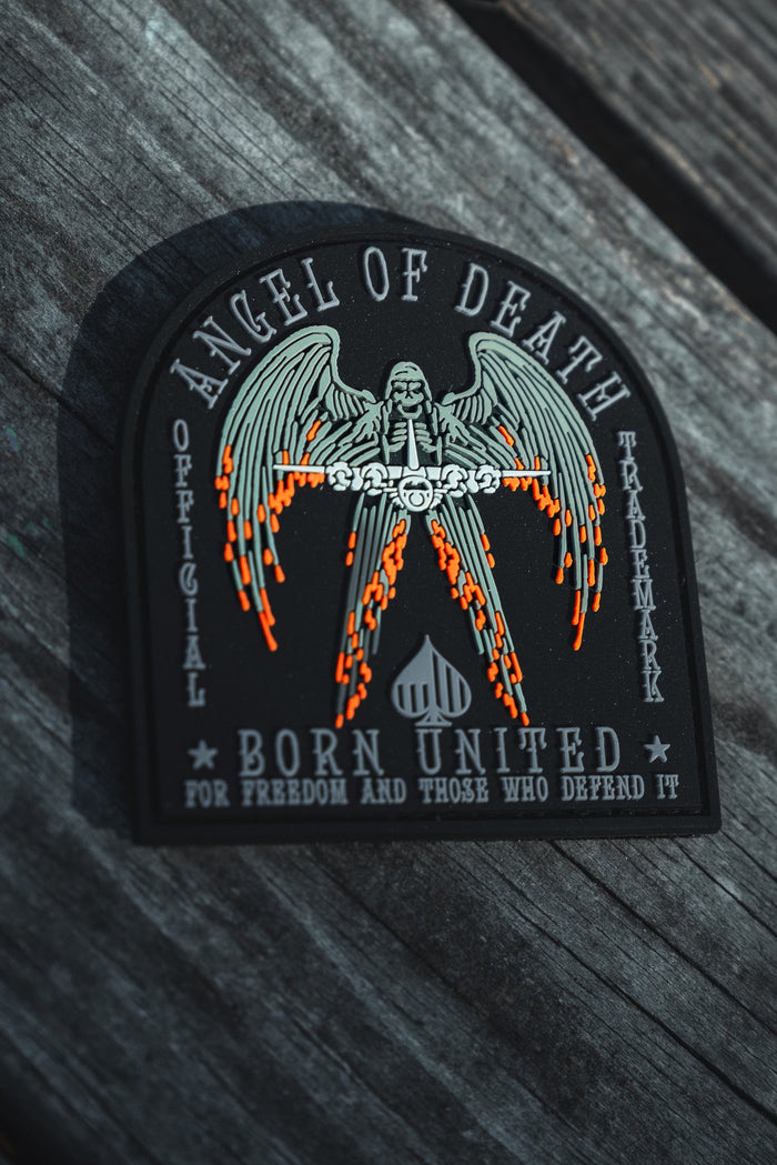 The Angel Of Death PVC Patch by Born United Private Label features an angel with flaming swords and spread wings. Text reads: Angel of Death, Born United, and For Freedom and Those Who Defend It. Displayed on a wooden background, it symbolizes courage.