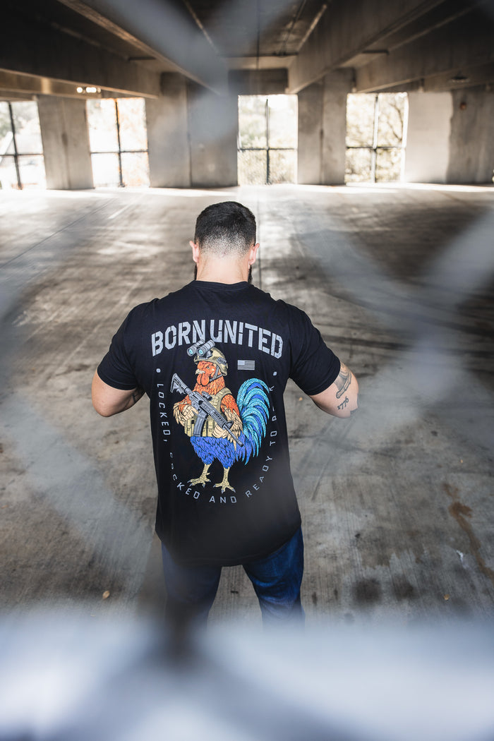 A man stands in an empty parking garage, his back to the camera, wearing the Born United Private Label "Battle Bird" T-shirt—a black 60/40 blend featuring a vibrant rooster graphic and BORN UNITED above it—shot through a blurred fence.
