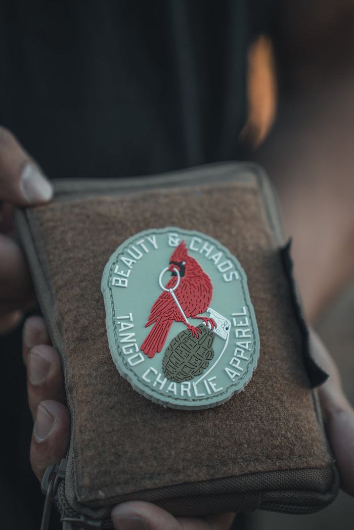 In a close-up, someone holds a pouch featuring the durable Beauty and Chaos 2.0 PVC Patch from Tango Charlie Apparel, showcasing a red wing design with Beauty & Chaos. With hands partly visible and the background blurred, this patch adds both style and resilience.