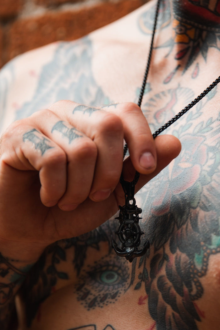 A close-up shows a person holding the Blackout Anchors Aweigh Necklace by Relentless Betrayal, attached to a stainless steel chain. Their tattooed chest and hands feature wings and floral patterns. This handcrafted piece offers elegance and includes a lifetime warranty.