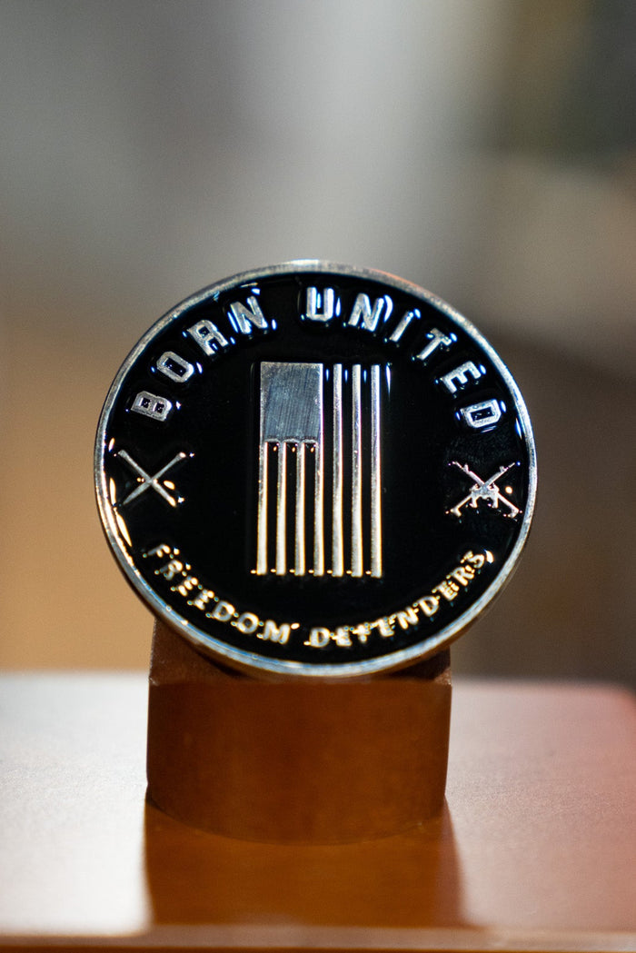 Display the Blue Line Flag Challenge Coin by Born United Private Labelโa round metal coin with a U.S. flag, crossed rifles, and the words BORN UNITED and FREEDOM DIVIDEDโupright on its wooden stand to show your support.