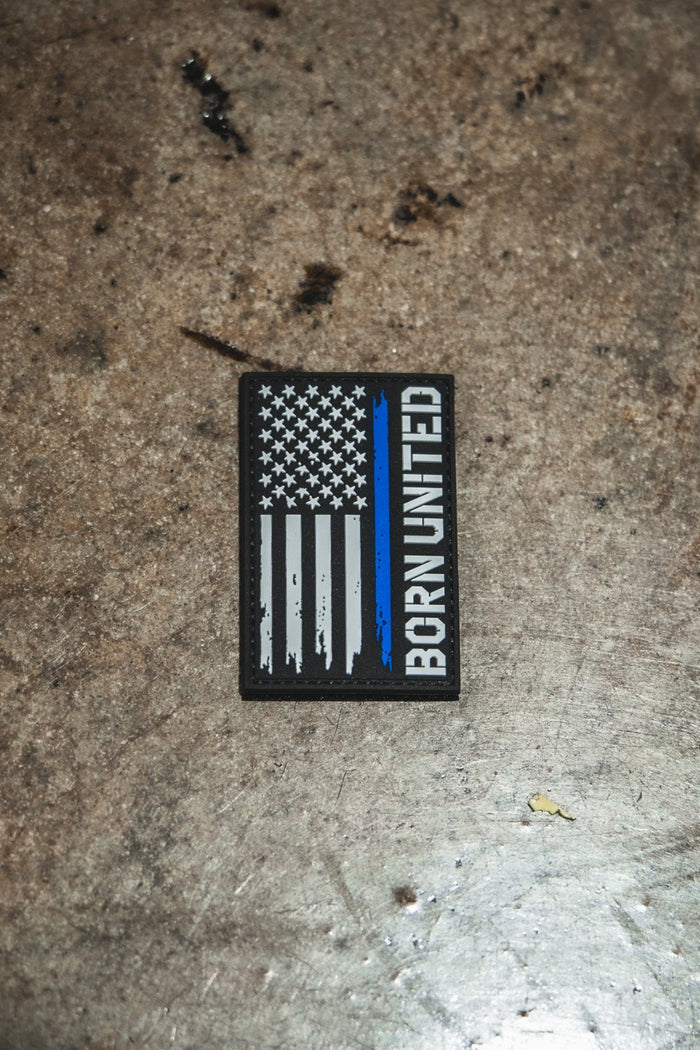 The Blue Line Flag PVC Patch by Born United Private Label features a black, white, and blue American flag with BORN UNITED written vertically on the right side, displayed on a textured surface.