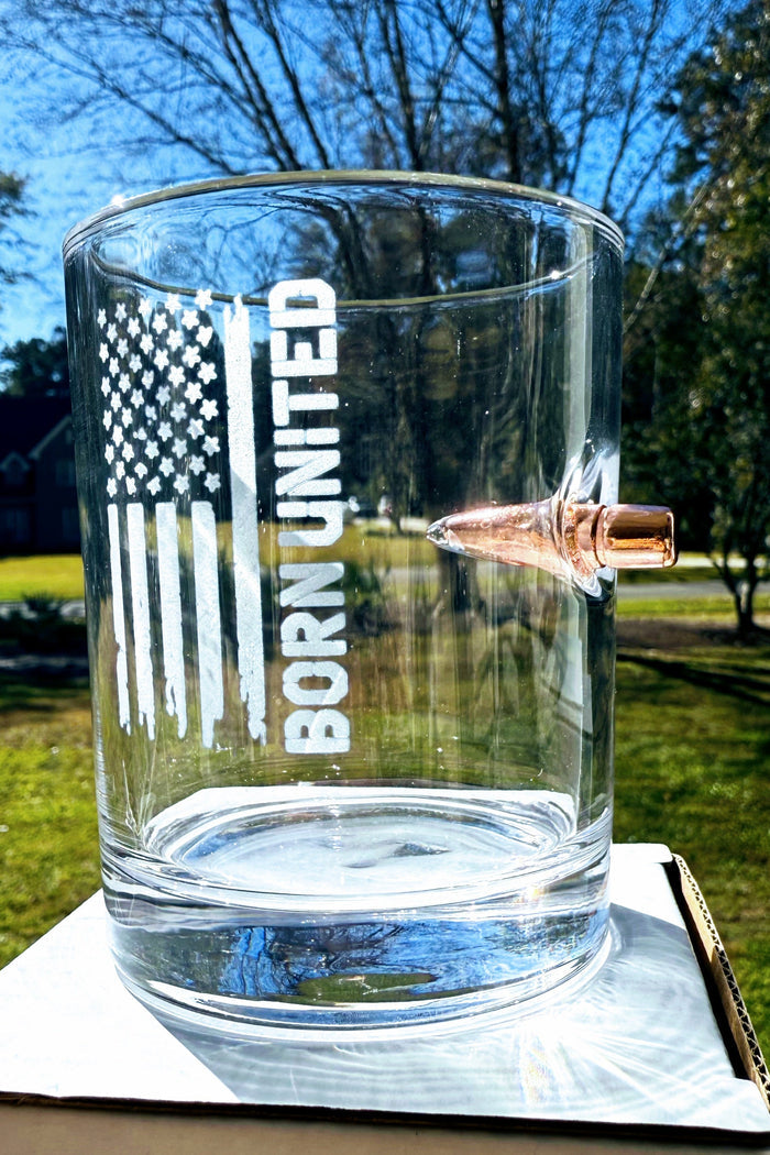 The Born United Bullet Whiskey Glass by Born United Private Label features a distressed American flag graphic with "BORN UNITED" etched on clear glass, plus a copper bullet embedded in the side.