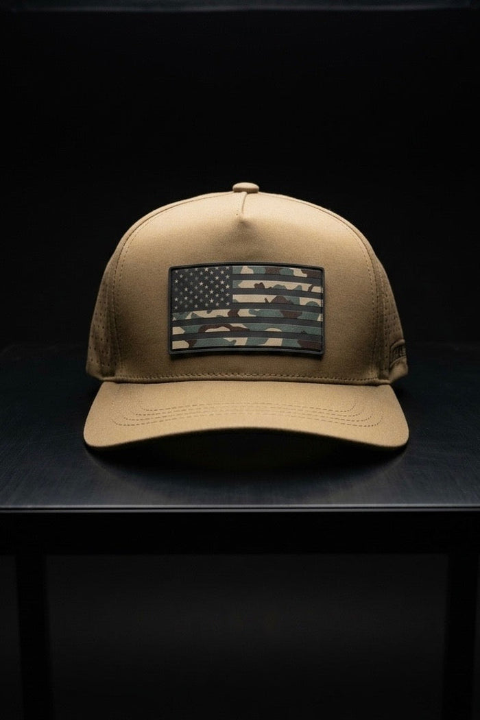 The Born United Private Label Premium Camo Flag Snapback rests on a dark surface, its front patch displaying the American flag in a green and brown camo pattern, highlighted against a shadowy background.