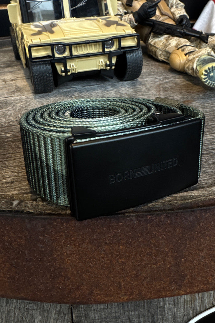 The BU Logo Belt - Camo by Born United Private Label, with green and black stripes and a rectangular buckle, is displayed on wood. A toy military vehicle and a camo-clad action figure complete the rugged scene.