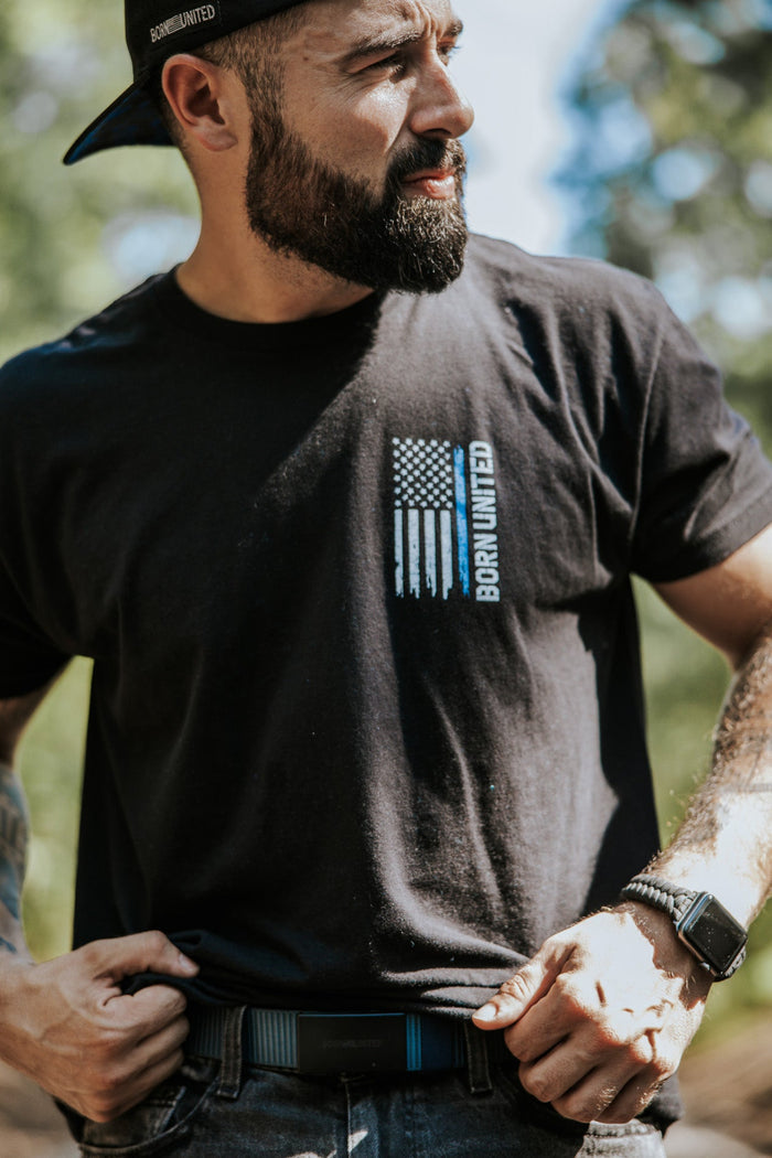 A bearded man outdoors in sunlight, wearing a black “Born United” tee with an American flag print, adjusts his Born United Private Label BU Logo Belt - Navy. He has tattooed arms, a smartwatch, and wears his black cap backwards.