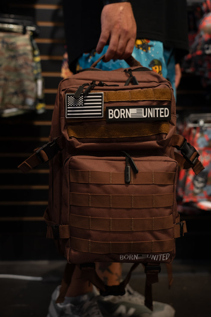 A person holds the Chocolate Tac-Pac - 25L by Born United Private Label—a brown tactical backpack with an American flag patch and a BORN UNITED patch, featuring organized storage—while standing in front of a display of colorful backpacks.