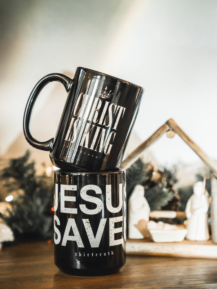Two black 13th โChrist is Kingโ mugs are stacked, one reading โCHRIST IS KING,โ the other โJESUS SAVES.โ Behind them, a nativity scene with festive greenery highlights the faith-filled message of these Christ is King Mugs.