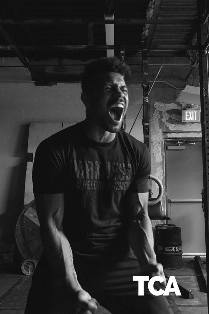 A man in a gym grips equipment, muscles straining with effort. The industrial setting features an exit sign and he wears Tango Charlie Apparel’s "Darkness is a Hell of a Coach" tee—engineered with perfect stretch for intense workouts.