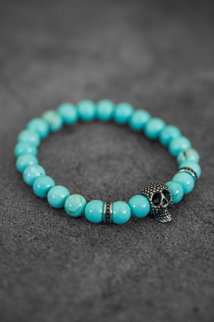 The Day of the Dead Bead Bracelet by Relentless Betrayal features turquoise beads, a metallic skull charm, and decorative spacers, shown here on a textured gray surface.