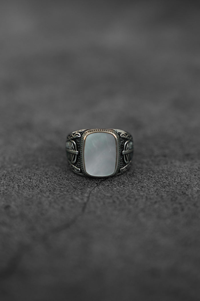 The Desire Opal Ring by Relentless Betrayal, featuring a large rectangular white opal and an ornate handcrafted band, is centered on a textured dark surface.