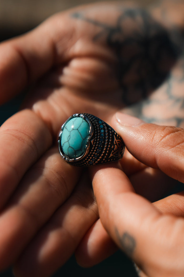 Close-up of hands holding a Relentless Betrayals Eye of the Storm Ring, styled with a large turquoise stone and detailed metalwork. The stainless steel highlights tattoos on the hands, while soft lighting creates shadows that emphasize the handcrafted jewelrys exquisite details.
