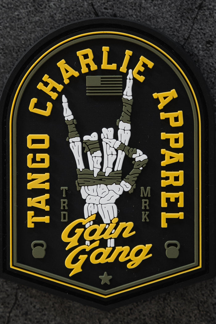 A 3-inch Velcro Gain Gang - Patch by Tango Charlie Apparel features a skeleton hand rock sign, yellow brand text, Gain Gang in script, small kettlebells, and an American flag on a gray textured background.