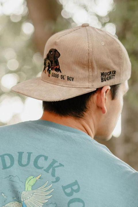 A man wears the Mucker Bucket Good Ol' Boy Tan Corduroy snapback hat with embroidered text and a dog image on the back, paired with a light blue-green shirt featuring a flying duck and “DUCK, BO,” creating a relaxed outdoor vibe.