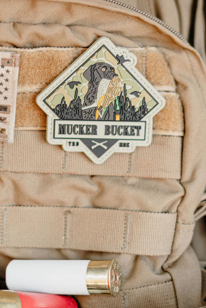 A Got One PVC Patch by Mucker Bucket, showing a dog with a stick, is attached to a tan tactical vest. Shotgun shells are lined along the bottom of the image.