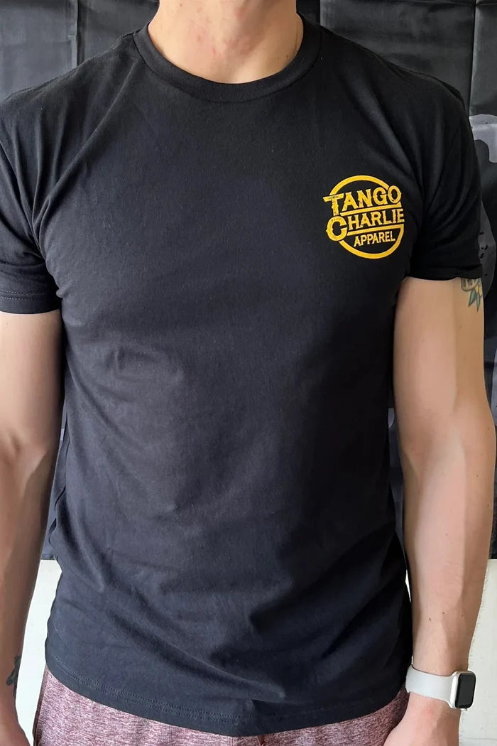 A fit individual wears a Tango Charlie Apparel Han Swolo T-Shirt in black with a yellow logo on the left chest, standing confidently before a black banner. Only their torso and part of the arms are visible.