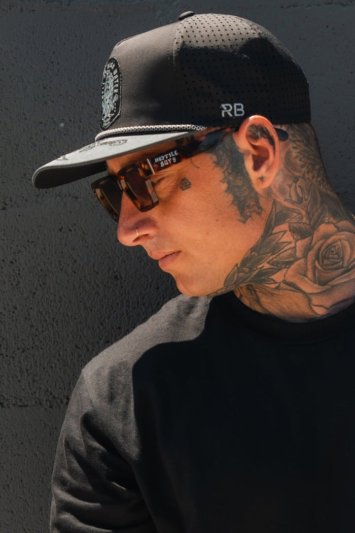 A tattooed individual wearing the Hell Or High Water Gray Snapback by Relentless Betrayal and sunglasses looks sideways. The rose and geometric tattoos on their neck and face reflect unyielding resilience, complemented by a black shirt against a gray backdrop.