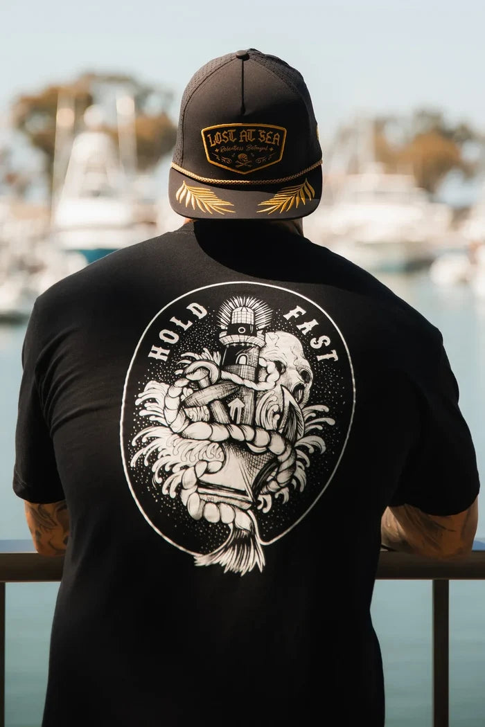A person in a black Relentless Betrayal Lost at Sea cap and a Hold Fast T-Shirt—Black leans on a railing, gazing over a marina with blurred boats and water in the background.