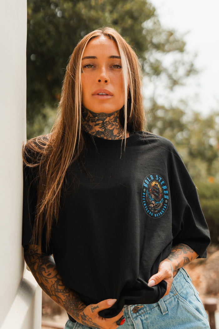 A woman with long brown hair and visible tattoos leans against an outdoor wall, wearing the Relentless Betrayal Hold Steady Blue - Premium Oversized Tee and light blue jeans, with blurred greenery in the background.