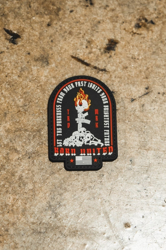 The Let The Darkness PVC Patch by Born United Private Label features a raised fist with a wrench, surrounded by text and โWork Unitedโ at the bottom in red, black, and white on a rugged brown surface.