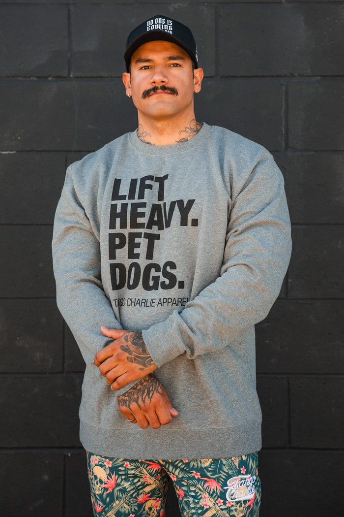 A man with a mustache stands against a black wall, wearing a black cap, colorful floral shorts, and the Lift Heavy. Pet Dogs Crewneck by Tango Charlie Apparel. He has hand tattoos and looks like someone who truly loves dogs.