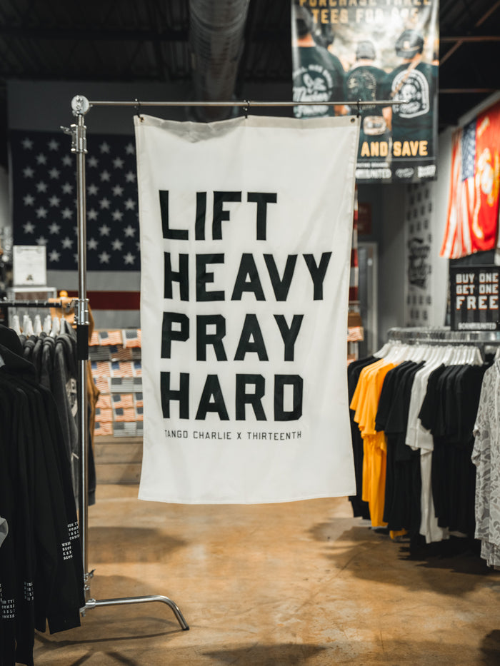 A Lift Heavy. Pray Hard. - Flag from Tango Charlie Apparel hangs in a clothing store, featuring bold text and surrounded by racks of shirts, patriotic decorations, and other gym-themed displays that celebrate faith and fitness.