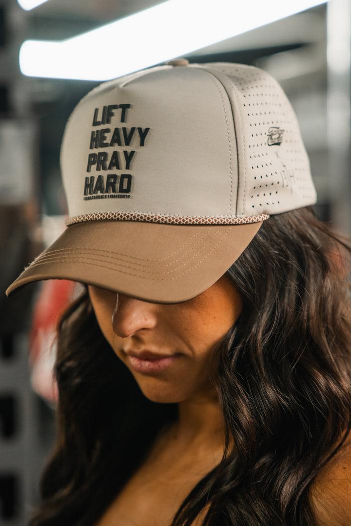 A woman with long, wavy brown hair wears the Tango Charlie Apparel Lift Heavy. Pray Hard. Hydro Snapback Hat in beige and tan, featuring bold black lettering on the front and a brim that partly shadows her face.