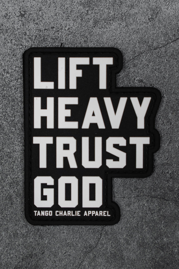The Lift Heavy. Trust God. - Patch by Tango Charlie Apparel is a black 3D PVC patch with bold white text, shown on a gray textured background.