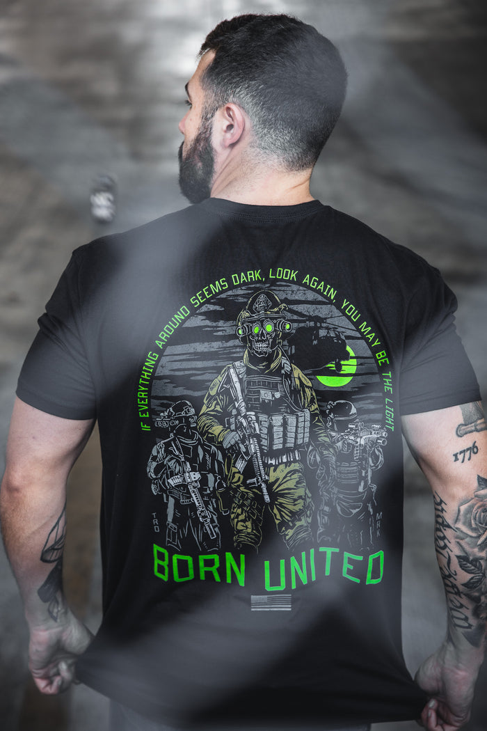 A man with short dark hair and a beard wears the Light in the Darkness t-shirt from Born United Private Label, featuring armored soldiers, neon green accents, and the phrase: You may be the Light in the Darkness.