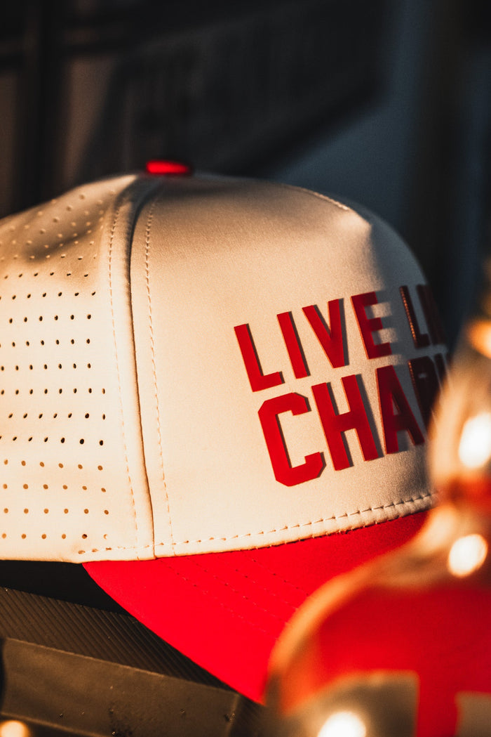 The Born United Private Label Live Like Charlie Ivory Snapback is a white and red 5-panel hat with bold red "LIVE LIKE CHARLIE" text, shown on a warmly lit surface. This breathable snapback adds a stylish touch.