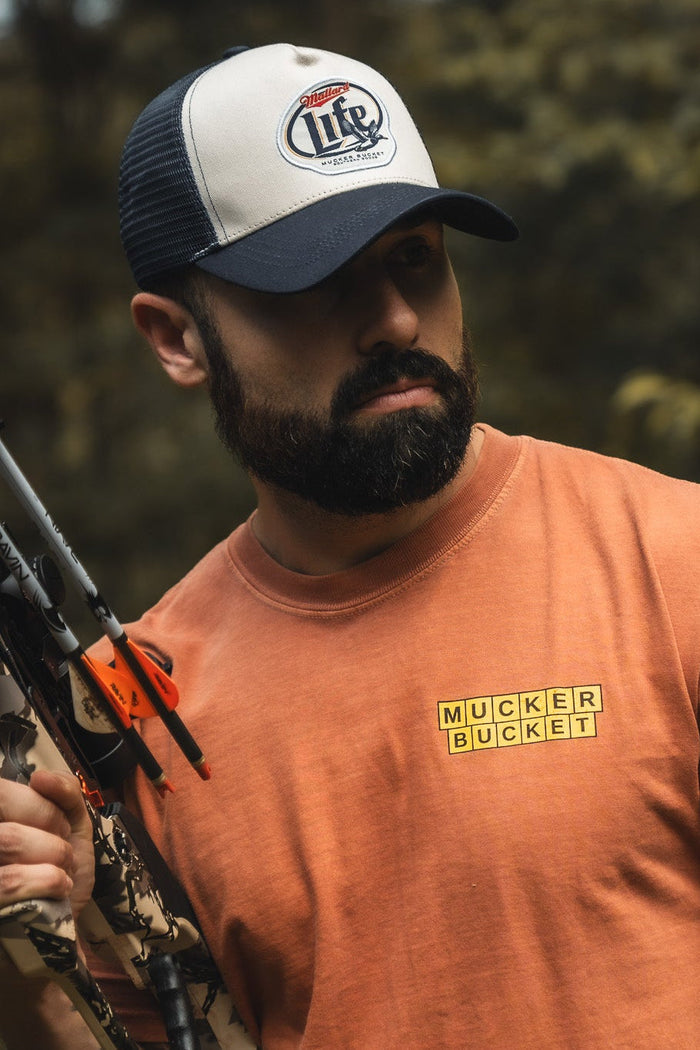 A bearded man in an orange Mucker Bucket t-shirt and a Mallard Life Trucker Bucket hat with mesh back holds a camo compound bow, standing outdoors among trees.