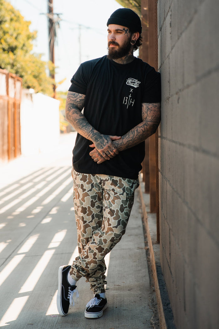 A tattooed man in a black beanie and tee stands outdoors, wearing Mucker Bucket Men's Classic Premium Joggers in Duck Camo and black sneakers, hands clasped, ready for running or relaxing.