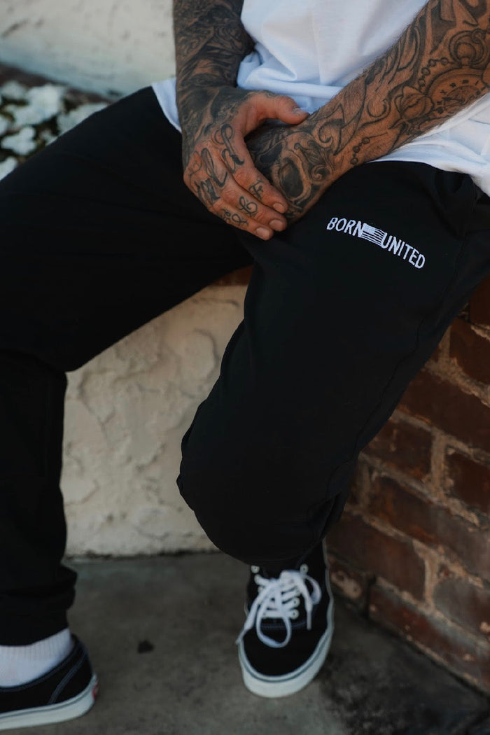 A person with tattooed hands sits on a brick ledge, wearing a white shirt, black-and-white sneakers, and Menโs Classic Premium Joggers | Stealth by Born United Private Label. Their hands are clasped over their lap.