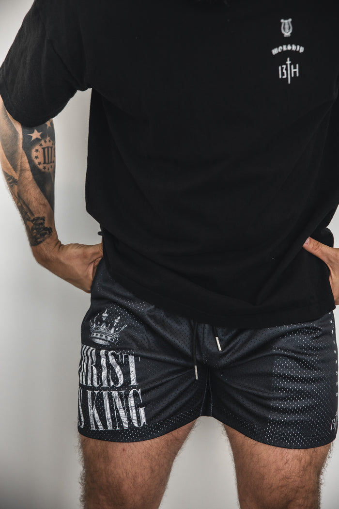 A person wears a black t-shirt and 13th Men's Mesh Lifestyle Shorts | 5.5" - Christ is King, with only the torso and upper legs visible. Their tattooed arms are also shown.