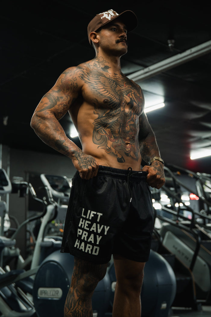 A muscular, tattooed man stands confidently in a gym wearing Tango Charlie Apparel's Men's Mesh Lifestyle Shorts | 5.5"- Lift Heavy Pray Hard Black, with exercise equipment visible in the background.
