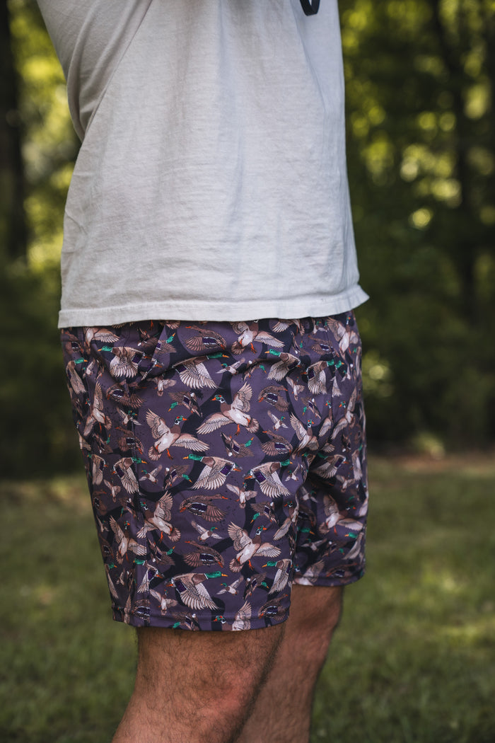 A person outdoors wears a white T-shirt and Mucker Bucket Men's Performance Athletic Shorts | 5.5"- Duck Wild, featuring a playful bird pattern. Blurred greenery in the background focuses attention on their torso and upper legs.