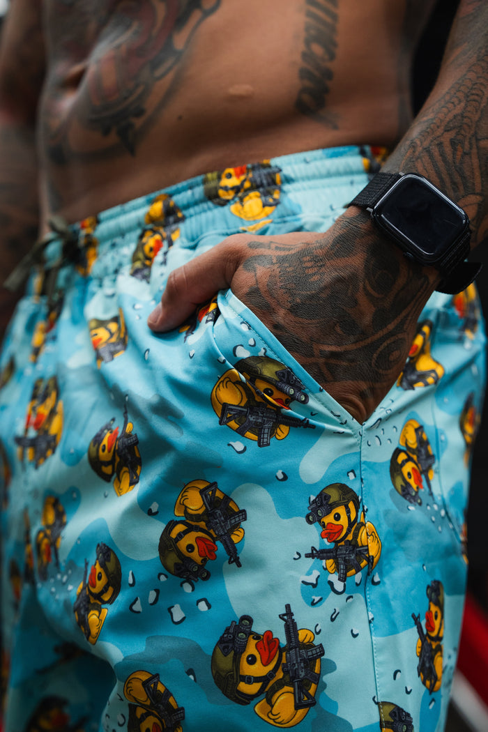 A close-up of a tattooed person wearing Born United Private Label’s Men's Performance Hybrid Shorts 5.5" | TactiQuack, featuring playful rubber duck soldiers. Their hand, with a smartwatch, rests in the pocket of these quick-dry shorts.