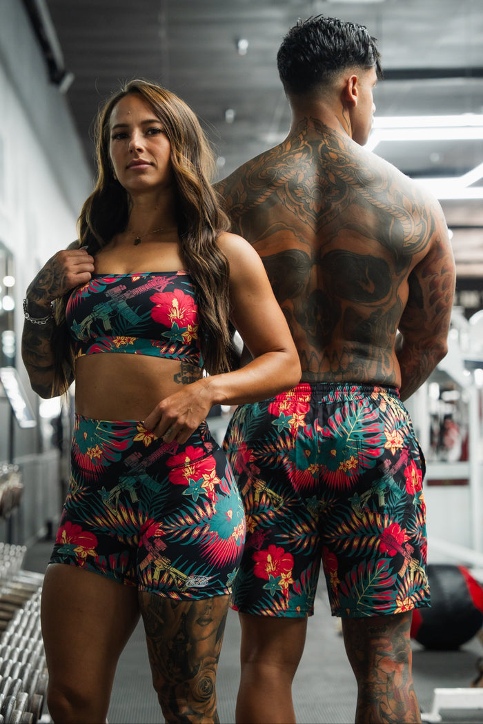 Two people with visible tattoos stand in a gym, wearing matching Tango Charlie Apparel outfits. The woman faces forward, while the man, back to the camera, sports Men's Performance Training Shorts | 5.5" - Tropical Assassin.