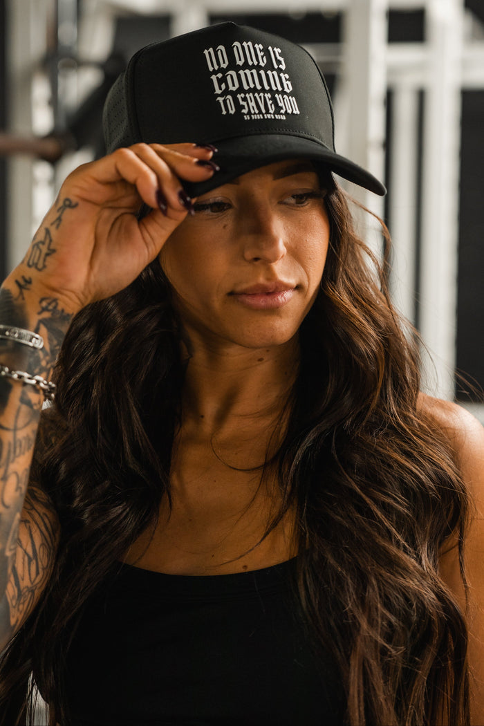 A woman with long dark hair and tattoos adjusts the Tango Charlie Apparel "No One Is Coming To Save You Hydro" snapback hat. The black, breathable cap features bold white text. She pairs it with a black tank top indoors, possibly at the gym.