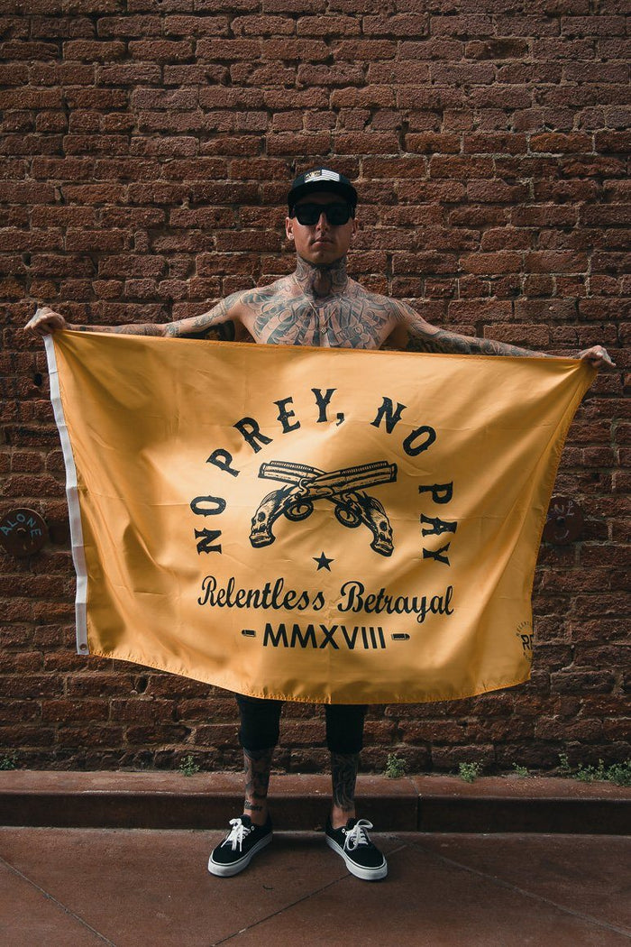 A tattooed person stands by a brick wall holding the No Prey, No Pay Flag by Relentless Betrayal, made from high-quality polyester. The flag features crossed pistols and No Prey, No Pay, Relentless Betrayal, MMXVIII. Theyre wearing a cap, sunglasses, tank top, shorts, and sneakers.