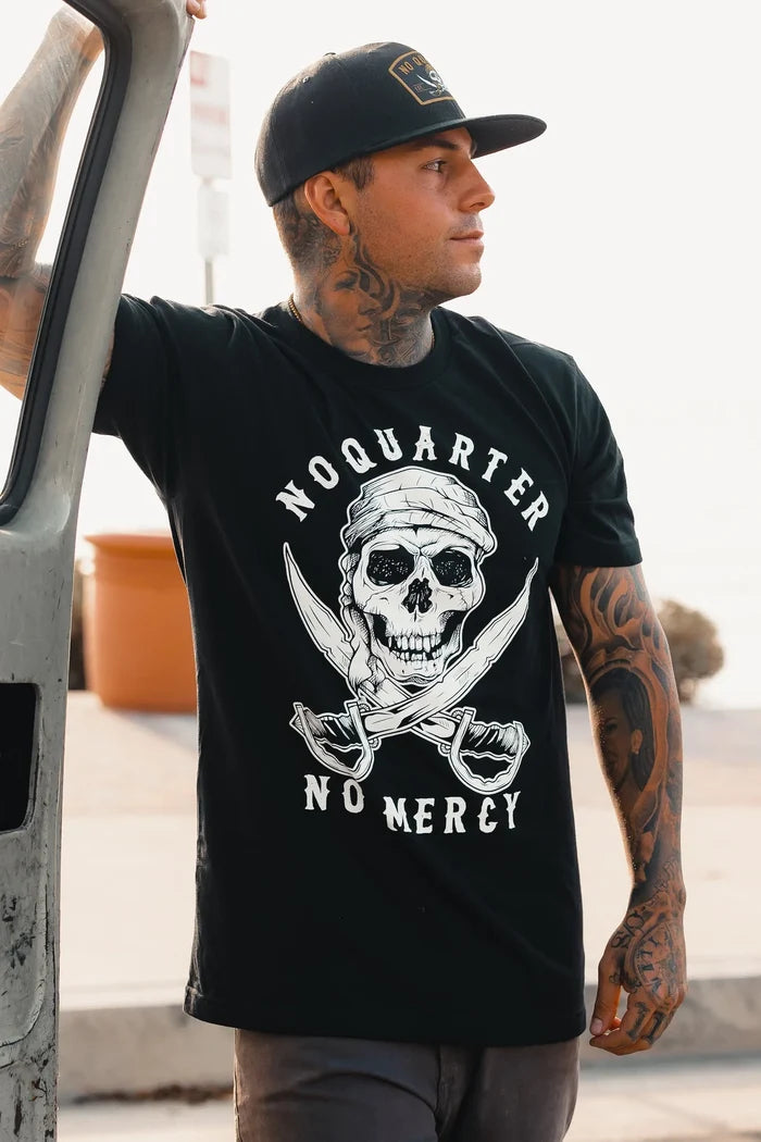 A tattooed man in a No Quarter Black T-Shirt by Relentless Betrayal, featuring a skull and crossed swords with No Quarter No Mercy, embodies mercilessness. He stands beside his vehicle, wearing a black cap, gazing aside against a blurred street and planter background.