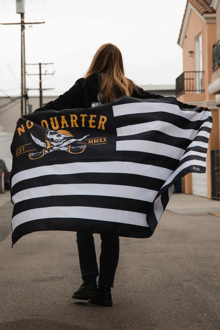 A person with long hair walks down a street holding the Relentless Betrayal No Quarter Stripe Flag V2 made of durable polyester, emblazoned with a skull and crossbones and No Quarter, fluttering against the overcast sky as buildings line their path.