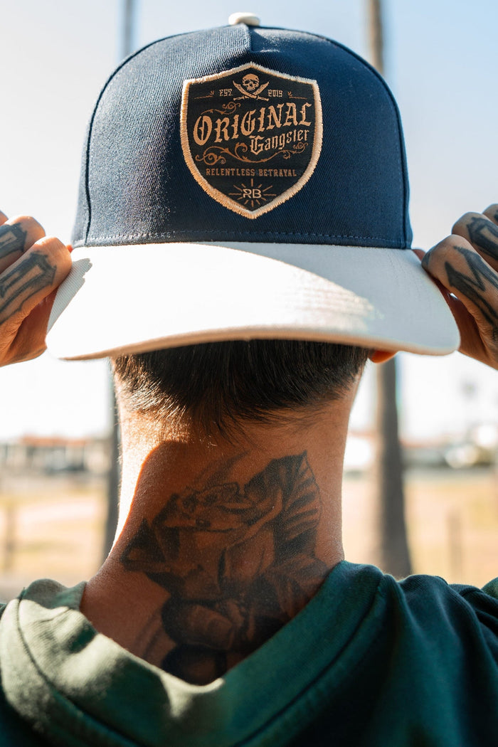 Amidst a sunny backdrop with palm trees, a person with tattooed hands and neck adjusts their OG Navy Snapback by Relentless Betrayal. The Original Gangster design adds an urban edge that complements their green shirt and enhances the scenes vibe.