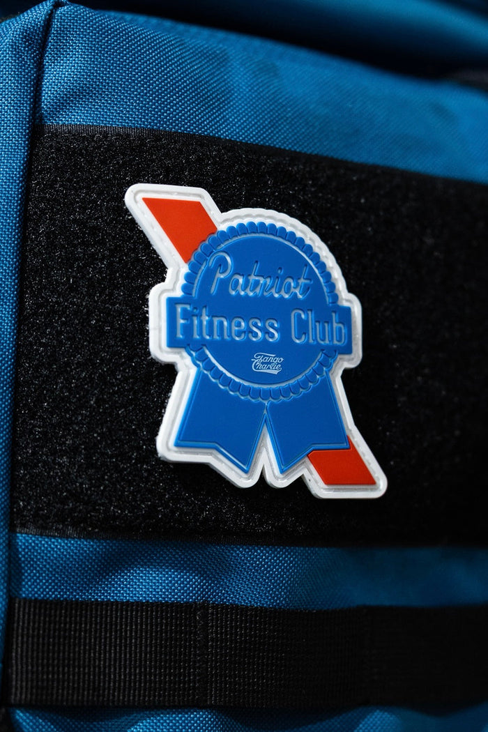 A Tango Charlie Apparel Patriots Fitness Club - Patch, shaped like a blue ribbon with red and white accents, is attached to the Velcro strip of a bright blue bag for a bold accessory.