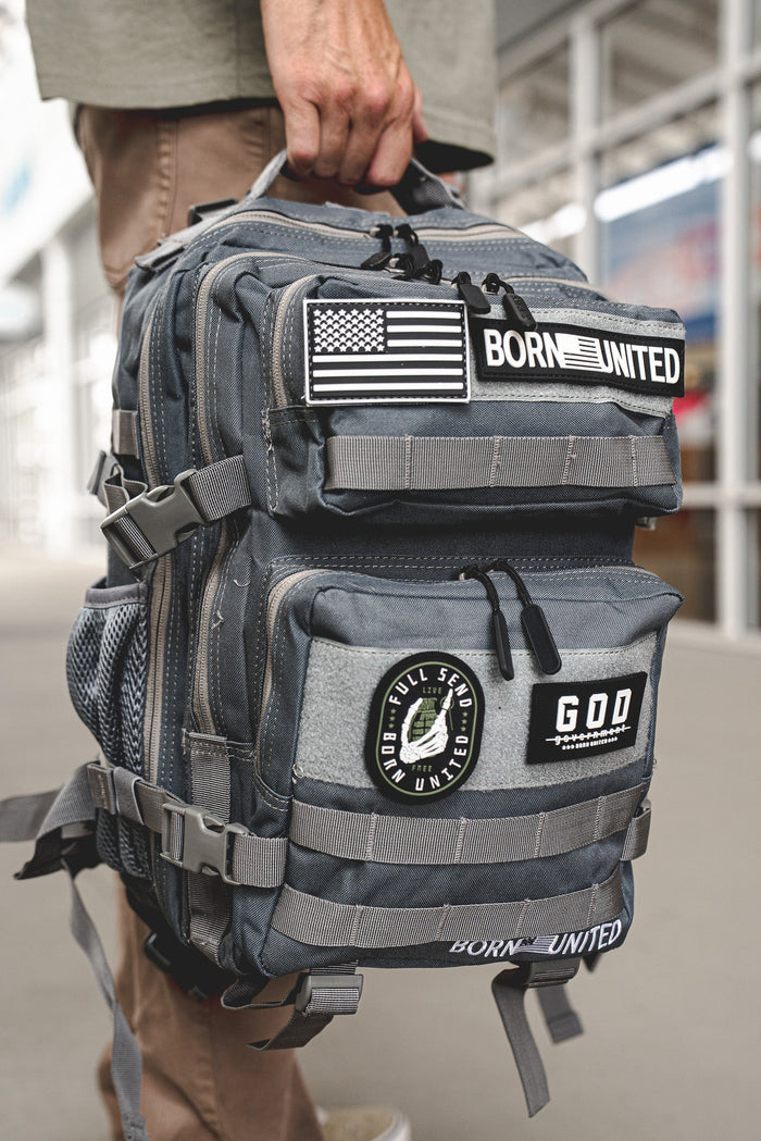 A person holds the Born United Private Label Standard Tac-Pac Ultimate Bundle backpack with patches—U.S. flag, BORN UNITED, and a GOD FULL SEND UNITED circle—while wearing a Born United tee outdoors in an urban area.