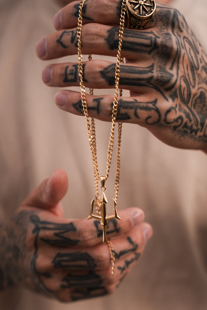 Tattooed hands clasp a Poseidons Trident Necklace by Relentless Betrayal, highlighting the maritime strength of Greek mythology jewelry. A ring with an intricate pattern adorns a finger against a softly blurred background, adding mystery.