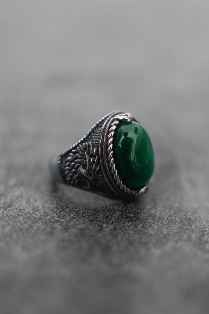 Relentless Betrayals Serpents Eye Ring, crafted in stainless steel, features intricate engravings and a central oval green gemstone that shimmers with an enchanting allure on a textured gray surface.