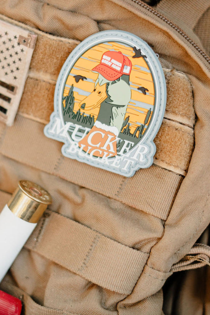 The Mucker Bucket Smart Duck PVC Patch, a 4x4 Velcro patch with a dog in a red cap against a sunset desert, is attached to a tan tactical vest beside a shotgun shell and an American flag patch.