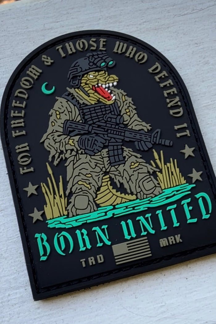 The TactiGator PVC Patch by Born United Private Label features an alligator in combat gear with rifle, the text "FOR FREEDOM & THOSE WHO DEFEND IT" and "BORN UNITED," blue water, grass, and a small American flag at the bottom.
